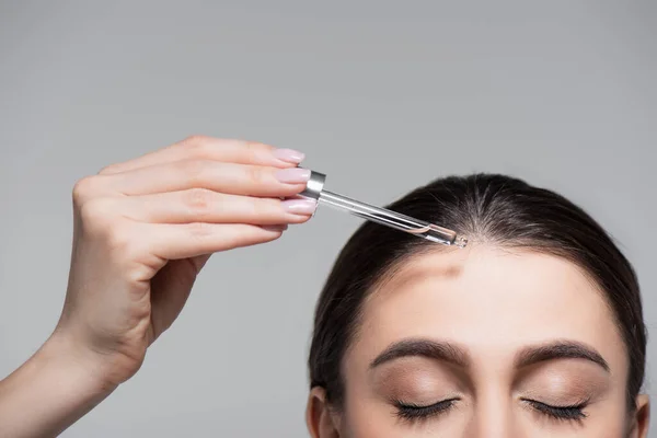 Vista cortada de jovem segurando pipeta com óleo perto de cabelo brilhante isolado em cinza — Fotografia de Stock