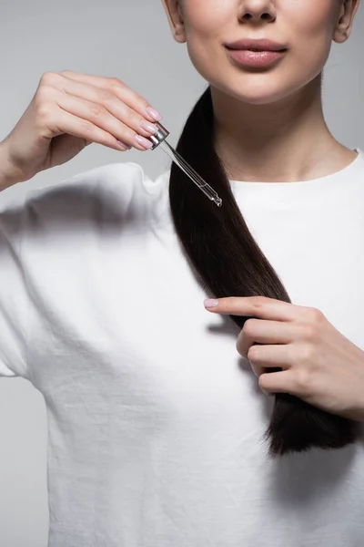Обрізаний вид молодої жінки в білій футболці, що тримає піпетку з маслом біля блискучого волосся ізольовані на сірому — стокове фото