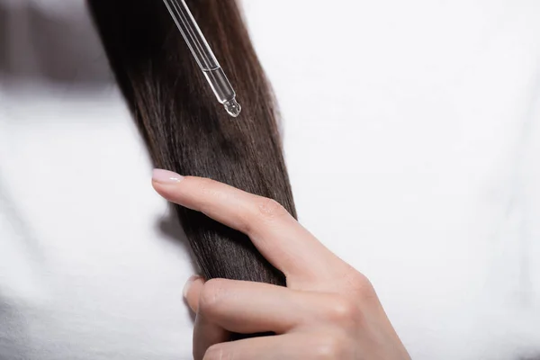 Vista recortada de la mujer en camiseta blanca sosteniendo la pipeta con aceite cerca del pelo brillante - foto de stock