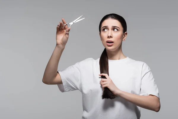 Shocked and brunette woman in white t-shirt holding scissors isolated on grey — Stock Photo