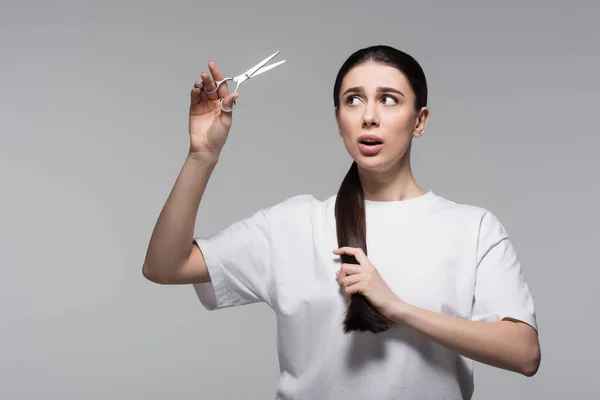 Femme choquée et brune en t-shirt blanc tenant des ciseaux isolés sur gris — Photo de stock