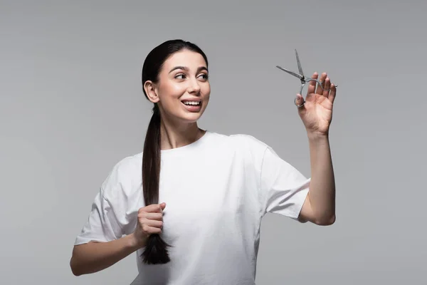 Весела жінка з блискучим волоссям тримає ножиці ізольовані на сірому — стокове фото