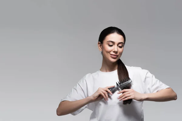 Cheerful woman in white t-shirt holding comb near straight hair isolated on grey — Stock Photo