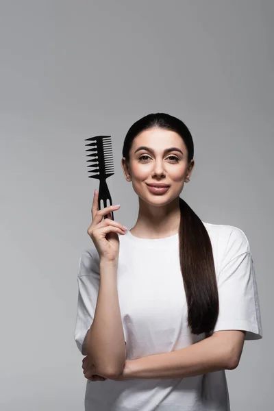 Happy woman in white t-shirt holding comb near straight hair isolated on grey — Stock Photo