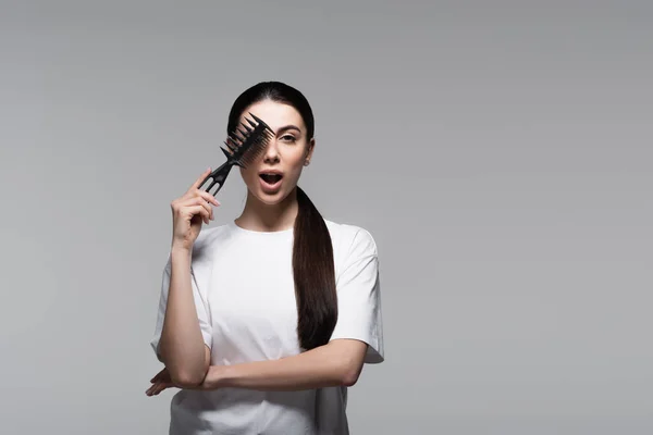 Young woman with open mouth holding comb near straight hair isolated on grey — Stock Photo