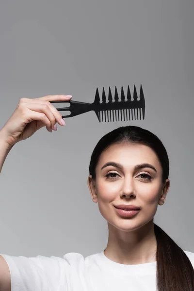 Cheerful woman in white t-shirt holding comb above straight hair isolated on grey — Stock Photo