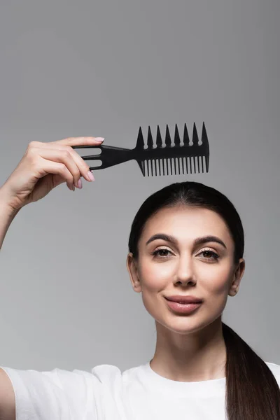 Femme gaie en t-shirt blanc tenant peigne au-dessus des cheveux raides isolés sur gris — Photo de stock