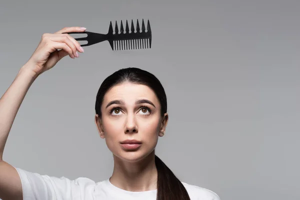 Mulher triste em t-shirt branca segurando pente acima do cabelo liso isolado em cinza — Fotografia de Stock