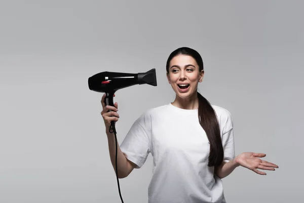 Mulher animada em t-shirt branca segurando secador de cabelo isolado em cinza — Fotografia de Stock