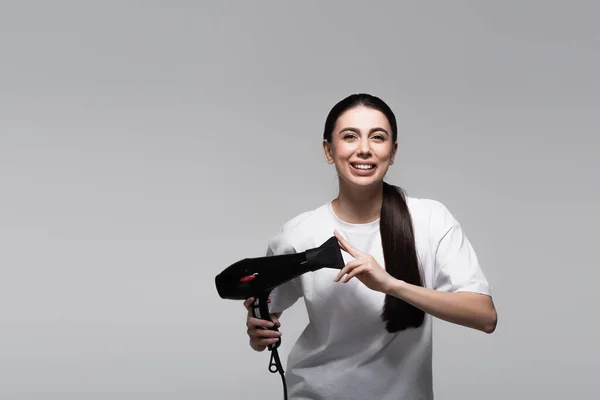 Mujer alegre en camiseta blanca sosteniendo secador de pelo aislado en gris - foto de stock