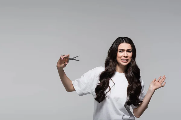 Незадоволена жінка з довгим хвилястим волоссям тримає ножиці ізольовані на сірому — стокове фото