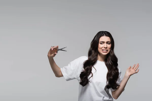 Mujer disgustada con el pelo ondulado largo sosteniendo tijeras aisladas en gris - foto de stock