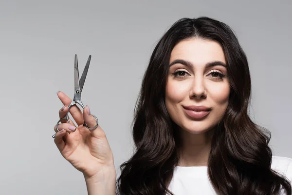 Donna sorridente con i capelli ondulati tenendo forbici isolate sul grigio — Foto stock