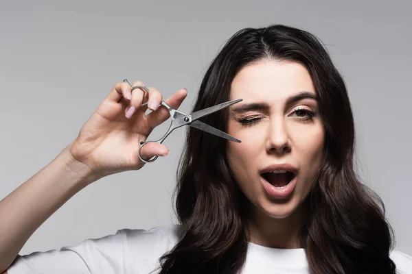 Woman with open mouth and wavy hair holding scissors isolated on grey — Stock Photo