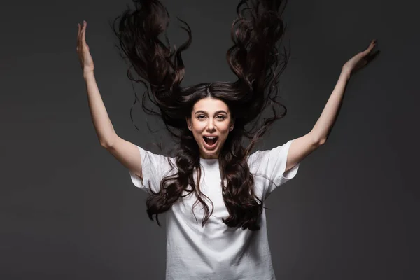 Mujer joven y sorprendida con el pelo ondulado gestos en gris oscuro - foto de stock