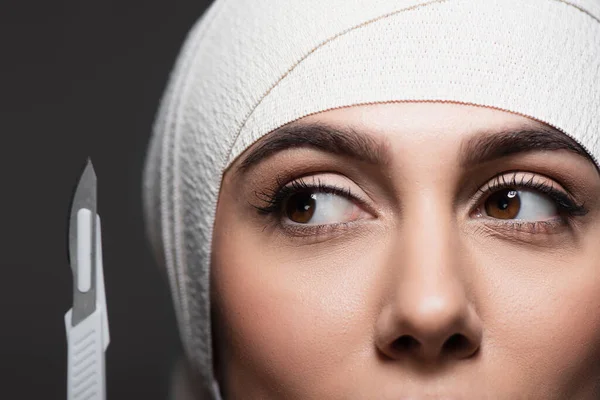 Vista recortada de la mujer en vendaje elástico en la cabeza cerca de cuchillo quirúrgico afilado aislado en gris - foto de stock