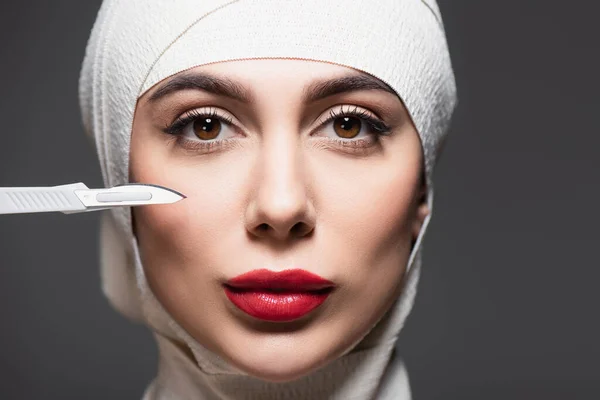 Gros plan de la femme en bandage élastique sur la tête près d'un couteau chirurgical tranchant isolé sur du gris — Photo de stock