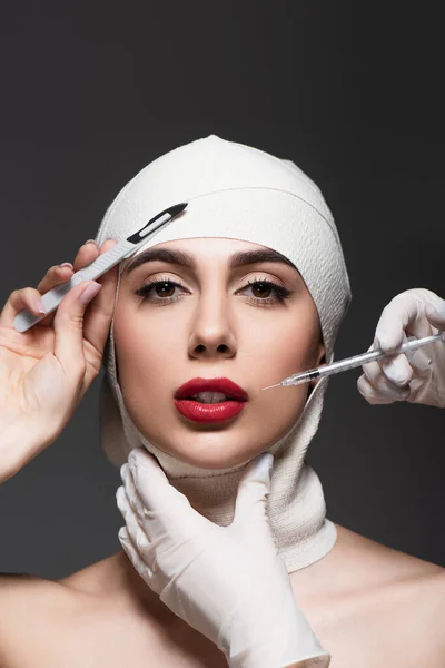 Plastic surgeons holding surgical knife and syringe near woman in elastic bandage on head isolated on dark grey — Stock Photo