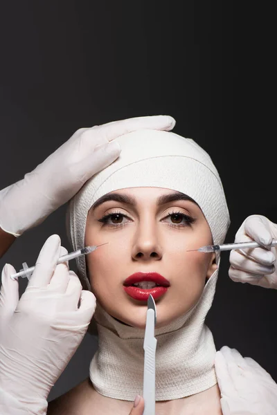 Cirurgiões plásticos em luvas de látex segurando faca cirúrgica e seringas perto da mulher em bandagem elástica na cabeça isolada em cinza escuro — Fotografia de Stock