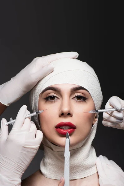 Cirujanos plásticos en guantes de látex que sostienen cuchillo quirúrgico y jeringas cerca de la mujer en vendaje elástico en la cabeza aislada en gris oscuro - foto de stock