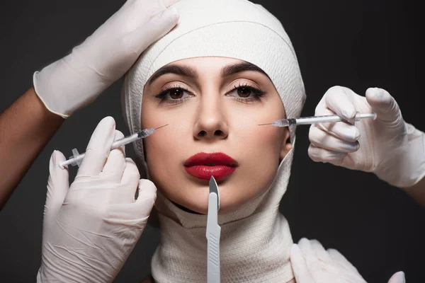 Cirurgiões plásticos segurando faca cirúrgica e seringas perto do paciente em bandagem elástica na cabeça isolada em cinza escuro — Fotografia de Stock