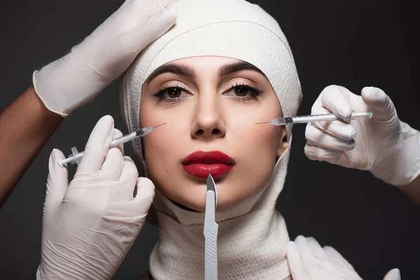 Plastic surgeons holding surgical knife and syringes near patient in elastic bandage on head isolated on dark grey — Stock Photo