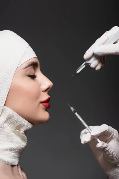 Vue latérale de la femme en bandage élastique sur la tête près des chirurgiens plasticiens tenant le couteau et la seringue chirurgicaux isolés sur gris foncé — Photo de stock