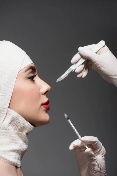 Vue latérale du patient en bandage élastique près des chirurgiens plasticiens tenant un couteau et une seringue chirurgicaux isolés sur du gris foncé — Photo de stock