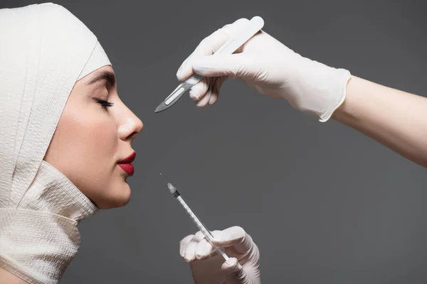 Side view of patient in elastic bandage near doctors holding surgical knife and syringe isolated on dark grey — Stock Photo