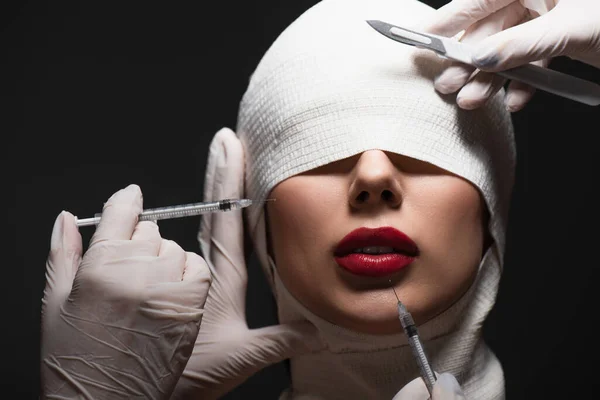 Femme en bandage élastique près des chirurgiens plasticiens avec couteau chirurgical et seringues isolées sur gris — Photo de stock