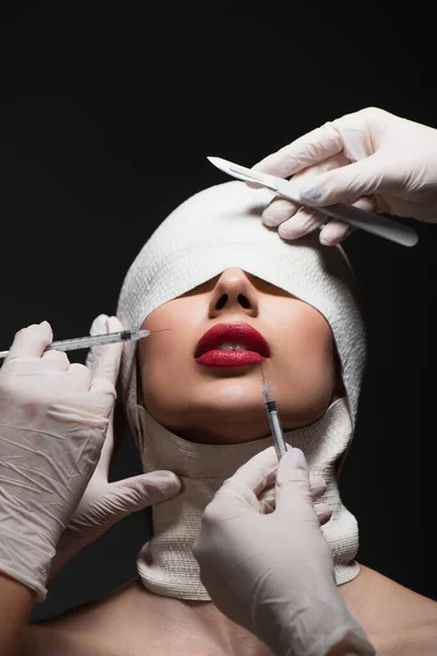Giovane paziente in fasciatura elastica vicino a chirurghi plastici con coltello chirurgico e siringhe isolate su grigio — Foto stock