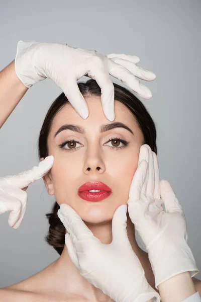 Plastic surgeons in latex gloves touching face of young woman isolated on grey — Stock Photo