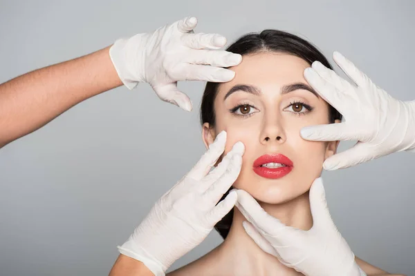 Пластические хирурги в латексных перчатках трогают лицо брюнетки женщины, изолированной на серой — стоковое фото