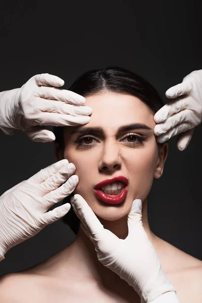 Cirujanos plásticos en guantes de látex tocando la cara de la joven morena aislada en negro - foto de stock