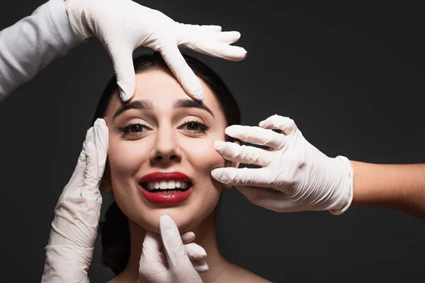 Cirujanos plásticos en guantes de látex tocando la cara de la joven sonriente aislada en negro - foto de stock