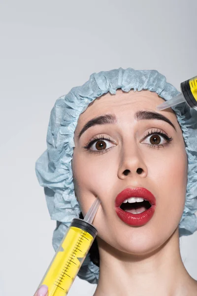 Huge syringes near face of shocked young woman in surgical cap isolated on grey — Stock Photo