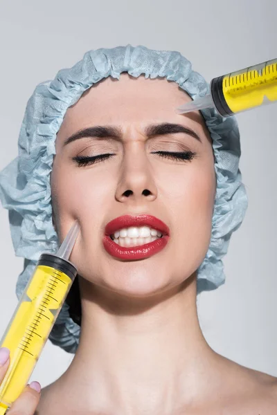 Огромные шприцы возле лица напуганной молодой женщины в хирургической шапке, изолированной на сером — стоковое фото