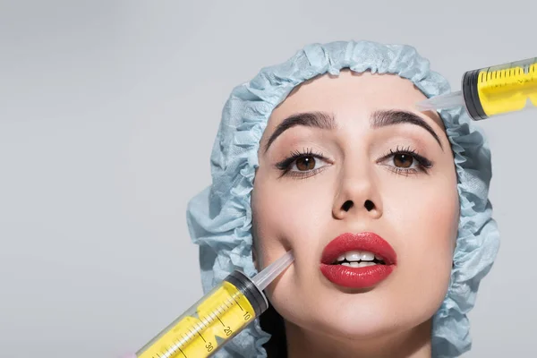 Huge syringes near face of young woman with red lips and surgical cap isolated on grey — Stock Photo