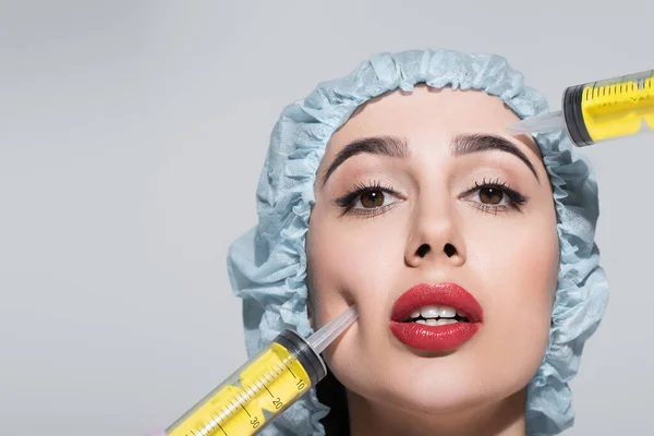 Seringues énormes près du visage de la jeune femme avec lèvres rouges et capuchon chirurgical isolé sur gris — Photo de stock