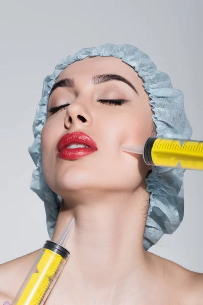 Enormes jeringas cerca de la cara de la mujer joven en la tapa quirúrgica aislado en gris - foto de stock
