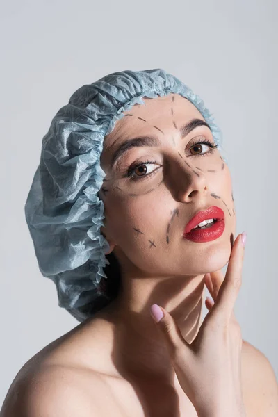 Молодая женщина в синей хирургической кепке с голыми плечами и отмеченными линиями на лице, изолированном на сером — стоковое фото