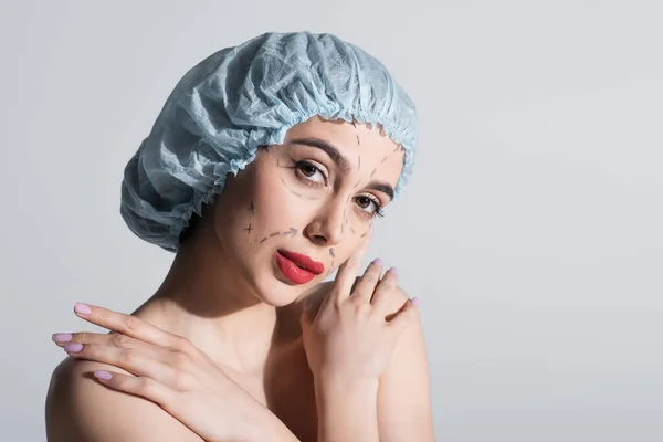 Jeune femme en bonnet chirurgical bleu avec des lignes marquées sur le visage embrassant les épaules nues isolé sur gris — Photo de stock