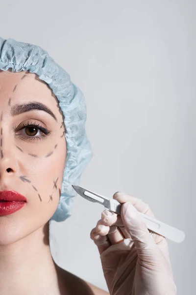 Обрезанный вид пластического хирурга в латексной перчатке, держащего скальпель рядом с женщиной с отмеченными линиями на лице, изолированными на сером — стоковое фото