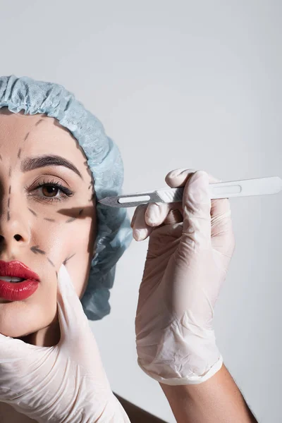 Частичный вид пластического хирурга в латексных перчатках, держащего скальпель рядом с женщиной с отмеченными линиями на лице, изолированными на сером — стоковое фото