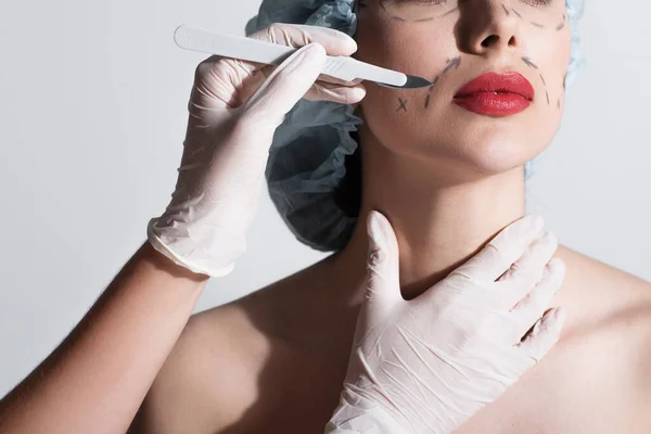 Visão recortada de cirurgião plástico em luvas de látex segurando bisturi perto de lábios vermelhos de mulher com linhas marcadas no rosto isolado em cinza — Fotografia de Stock