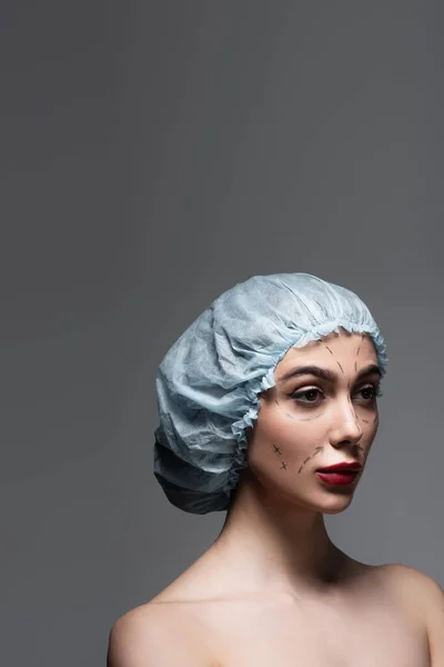 Mujer joven en gorra quirúrgica con líneas marcadas en la cara mirando hacia otro lado aislado en gris oscuro - foto de stock