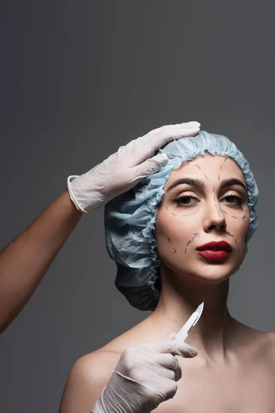 Plastic surgeon in latex gloves holding scalpel near woman in surgical cap with marked lines on face isolated on dark grey — Stock Photo