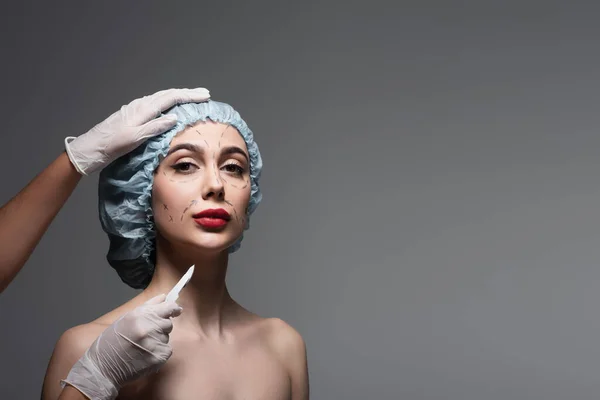 Chirurgien plasticien en gants de latex tenant le scalpel près de la femme avec des lignes marquées sur le visage et les épaules nues isolées sur le gris foncé — Photo de stock