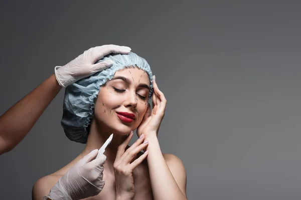 Chirurgo plastico che tiene il bisturi vicino a donna soddisfatta in berretto chirurgico con linee marcate sul viso isolate su grigio scuro — Foto stock