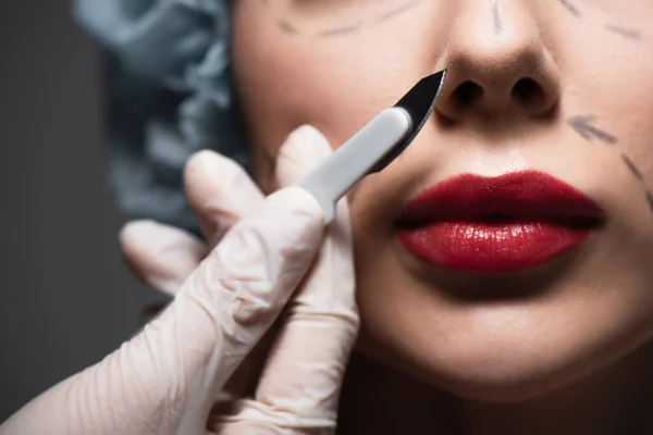 Close up of plastic surgeon in latex glove holding scalpel near woman with marked lines on face isolated on grey — Stock Photo